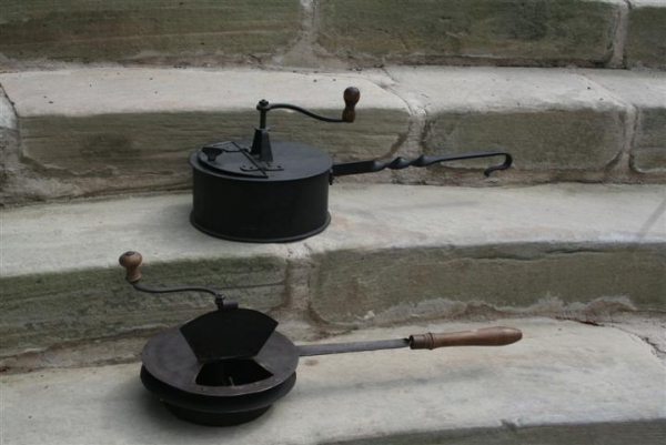 Die beiden in unserem Museum ausgestellten Röstpfannen aus Eisenblech mit integriertem Rührwerk stammen aus der Zeit um 1900. Der Kurbelmechanismus setzt einen Rührstab in Gang, der die Kaffeebohnen am Boden des Gerätes bewegt, so dass sie nicht anbrennen und von allen Seiten gleichmäßig geröstet werden. Für den Röstvorgang stellte man die Pfannen in die offene Glut einer Feuerstelle oder eines Ofens, beziehungsweise in die Ringöffnung eines Kohleherdes. Die grünlich-gelben Rohbohnen mussten so lange erhitzt werden, bis sie aufbrachen, sich um ein Drittel vergrößerten und eine braune Farbe annahmen. Während dieses Prozesses entfalteten sie ihr charakteristisches Kaffeearoma. Der Röstvorgang dauerte – je nach gewünschtem Bräunungsgrad - etwa zwanzig Minuten. Danach mussten die Bohnen auskühlen, bevor sie in einer Handmühle gemahlen und schließlich aufgebrüht werden konnten.  